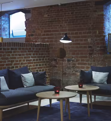 Mysig lounge med synliga tegelväggar, två blå soffor med vita kuddar, två runda träbord och en golvlampa. Två små röda ljus på borden skapar en varm atmosfär. Stora fönster ger naturligt ljus.