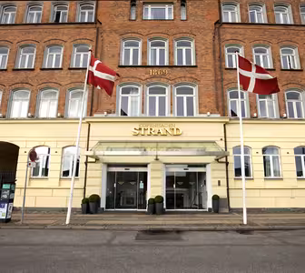 Bilden visar entrén till Copenhagen Strand hotel, en tegel- och gulmålad byggnad med två danska flaggor som vajar ovanför glasdörrarna. Byggnadens fasad har rader av fönster och årtalet 1869 inskrivet ovan.