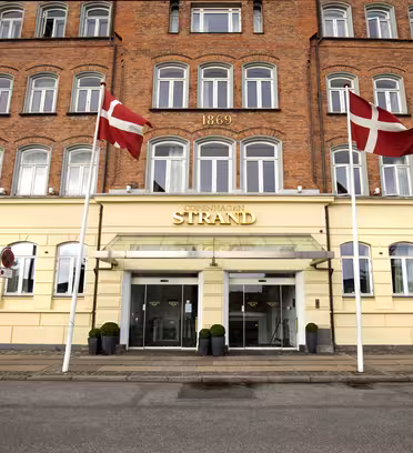 Bilden visar entrén till Copenhagen Strand hotel, en tegel- och gulmålad byggnad med två danska flaggor som vajar ovanför glasdörrarna. Byggnadens fasad har rader av fönster och årtalet 1869 inskrivet ovan.
