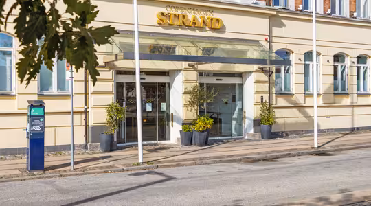 Entré till Copenhagen Strand hotel, med en gul fasad med stora glasdörrar, baldakin och krukväxter. En parkeringsautomat står på trottoaren, och flaggor syns på stolpar i närheten.