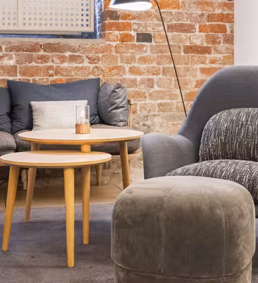 En mysig sittgrupp med en modern grå fåtölj, en mörkgrå vadderade ottoman, två runda sidobord i trä och en mörkgrå soffa mot en exponerad tegelvägg. En hög golvlampa ger värme till utrymmet.