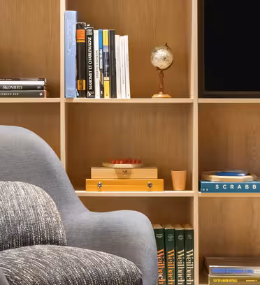 En mysig läshörna har en grå fåtölj med en mönstrad kudde, en bokhylla i trä fylld med böcker, en miniatyrglob, en Scrabble-spellåda och en liten TV-skärm i det övre högra hörnet.