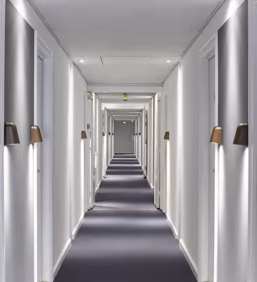 En lång, väl upplyst hotellhall med mörk matta, vita väggar och stängda dörrar på båda sidor. Varje dörr har en brun och vit lampett ovanför och ett dörrhandtag i mässing med nyckelkortslås. Korridoren verkar leda till en avlägsen utgång.