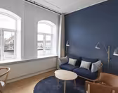 En modern vardagsrumsdel med en blå accentvägg, med en blå soffa med kuddar, ett runt soffbord i trä och en brun läderstol. Höga lampor står bredvid soffan. Stora fönster ger utsikt över vattnet och byggnader utanför.