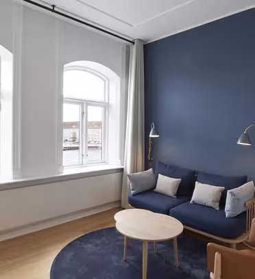 En modern vardagsrumsdel med en blå accentvägg, med en blå soffa med kuddar, ett runt soffbord i trä och en brun läderstol. Höga lampor står bredvid soffan. Stora fönster ger utsikt över vattnet och byggnader utanför.