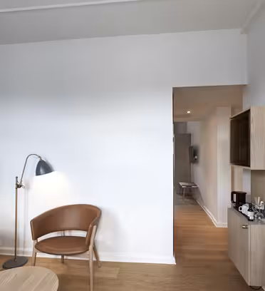 Ett modernt, minimalistiskt hotellrum har en blå och vit vägg, en trästol, ett litet runt bord, en golvlampa, en monterad TV och ett träskåp med en kaffestation längs en hall som leder till ett annat område.