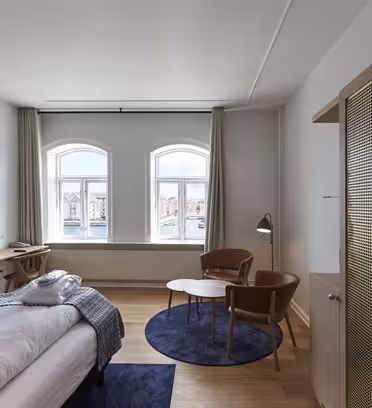 Hotellrum med stora fönster med utsikt över vattnet. I rummet finns en säng, TV, två bruna stolar, ett runt bord på en blå matta och en garderob i trä. Väggarna är vita med trägolv, och naturligt ljus lyser upp utrymmet.