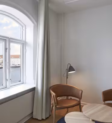 Ett litet rum med fönsterutsikt över vatten och byggnader. Två bruna läderstolar omger ett ljust cirkulärt bord i trä. En golvlampa med svart skärm är placerad mellan stolarna. Vita väggar och en gardin ramar in fönstret.