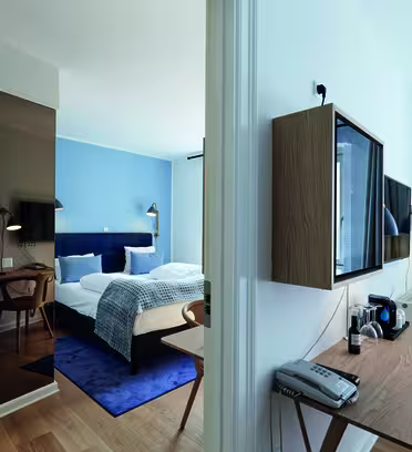 Ett modernt hotellrum med en stor säng, blå och vita sängkläder och en blå accentvägg. Det finns ett träskrivbord i förgrunden med telefon och kaffeinstallation. Rummet har ljusa trägolv och modern inredning.