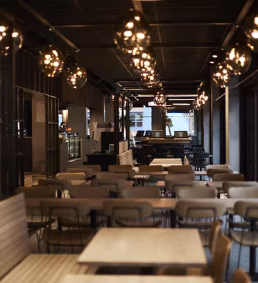 En modern, svagt upplyst restauranginteriör med träbord och stolar ordnade i rader. Hängande sfäriska taklampor lyser upp utrymmet. Bakgrunden har en disk, trädetaljer och stora fönster.