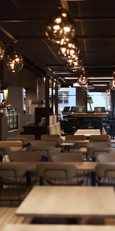 En modern, svagt upplyst restauranginteriör med träbord och stolar ordnade i rader. Hängande sfäriska taklampor lyser upp utrymmet. Bakgrunden har en disk, trädetaljer och stora fönster.