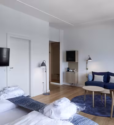 Modernt hotellrum med två snyggt bäddade sängar, en väggmonterad TV och en liten sittgrupp med en blå soffa, ett runt soffbord och en brun stol. Två stående lampor flankerar soffan. Ljusa trägolv kompletterar rummets minimalistiska design.