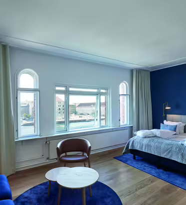 Ett modernt hotellrum med en vägg med blå accent har en dubbelsäng, en mysig sittgrupp med en blå soffa och brun stol samt stora fönster med utsikt över staden. Rummet har trägolv, ett runt soffbord och golvlampor.
