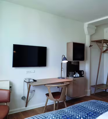 Minimalistiskt hotellrum med en monterad platt-TV, skrivbord i trä, moderna lampor, läderstol, öppen garderob och en snyggt bäddad säng med blåmönstrad släng. Rummet har ljusa trägolv och en vit vägg, vilket skapar en ren, mysig atmosfär.