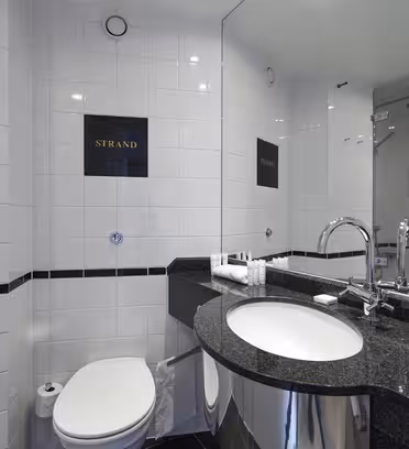 Ett modernt badrum med vita kaklade väggar har en snygg svart bänkskiva och inbyggt handfat. Takbelysning lyser upp området. En stor spegel reflekterar en duschkabin med en glasdörr. En toalett finns bredvid disken.