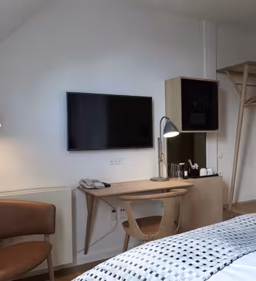 Ett modernt hotellrum har en snyggt bäddad säng med ett geometriskt mönstrat täcke. Mittemot sängen står en väggmonterad TV, ett träskrivbord med två lampor, en telefon och en kaffebryggare. En trästol och en öppen garderob finns också.