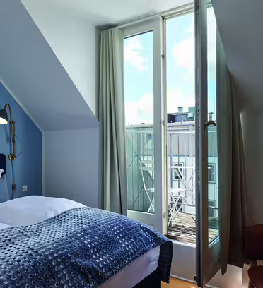 Ett mysigt sovrum med sluttande tak har en dubbelsäng med mönstrad filt, blå accentvägg och väggmonterad läslampa. Stora glasdörrar öppnar mot en liten balkong med utsikt över tak och moln. En brun stol sitter i hörnet.