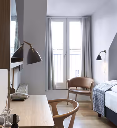 Ett modernt hotellrum med ett skrivbord och stol i trä, en säng med mönstrad filt och ett stort fönster med grå gardiner. Två svarta lampor är placerade på skrivbordet och vid sängen. En liten stol står nära fönstret.