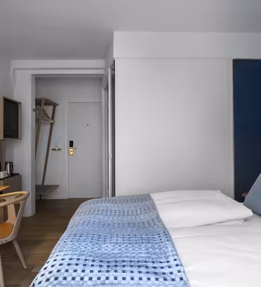 Modernt hotellrum med en mysig säng, blå och vita sängkläder och en vägg med marin accent. Till vänster finns ett skrivbord i trä med stol och lampa, och en liten öppen garderob och minibar syns nära entrén.
