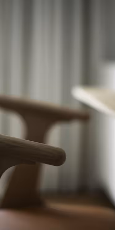 En närbild av en trästol med ett böjt ryggstöd bredvid ett träbord. En vas med gröna blad syns delvis på bordsskivan. Mjukt, naturligt ljus kommer in från ett fönster, vilket skapar en lugn och minimalistisk atmosfär.