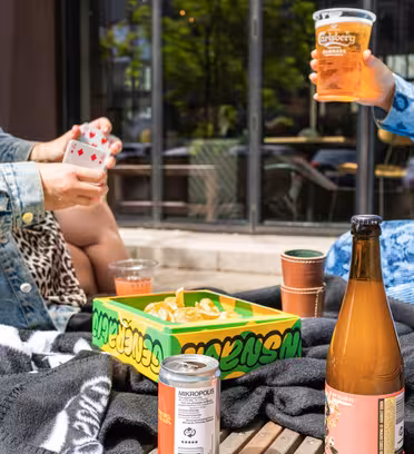 Två personer som sitter utomhus, en med spelkort. En Carlsberg-kopp och en flaska dryck står på ett bord med en burk och en snackslåda. Båda bär casual jackor. Bakgrunden visar ett fönster som reflekterar grönska.