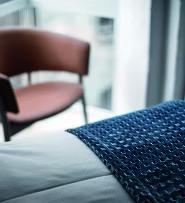 En suddig bakgrund av en brun stol vid ett fönster. I förgrunden, en säng med en texturerad blå släng draperad över vita sängkläder. Scenen framstår som lugn och modern.