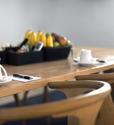 Ett konferensbord med trästolar är dukat med kaffekoppar, fat och pennor på anteckningsblock. En svart korg i mitten rymmer bananer och äpplen. Miljön är väl upplyst och arrangerad för ett möte eller diskussion.