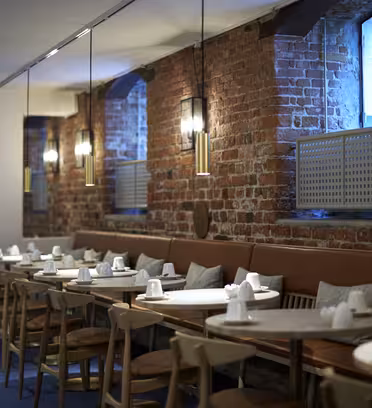 En mysig restauranginredning med exponerade tegelväggar och varm belysning. Träbord och stolar är snyggt arrangerade, varje set med koppar och servetter. Stora fönster släpper in naturligt ljus och skapar en välkomnande atmosfär.