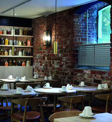 En mysig restaurang med träbord och stolar, med vita servetter. Tegelväggarna har ett stort fönster och hyllor fyllda med färgglada burkar med kryddor, vilket skapar en varm och inbjudande atmosfär.