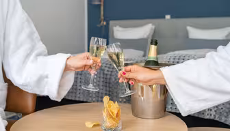 Två personer i vita dräkter skålar med champagneglas i ett modernt hotellrum. En hink med en champagneflaska och ett glas potatischips står på ett träbord mellan dem. Sängen, med vita kuddar, står i bakgrunden.