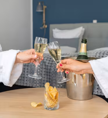 Två personer i vita dräkter skålar med champagneglas i ett modernt hotellrum. En hink med en champagneflaska och ett glas potatischips står på ett träbord mellan dem. Sängen, med vita kuddar, står i bakgrunden.