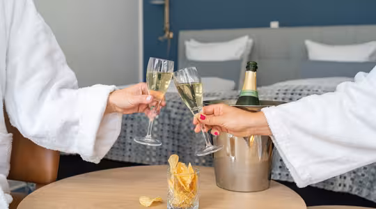 Två personer i vita dräkter skålar med champagneglas i ett modernt hotellrum. En hink med en champagneflaska och ett glas potatischips står på ett träbord mellan dem. Sängen, med vita kuddar, står i bakgrunden.