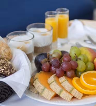 En frukostbricka med fullkornsrullar i en korg, en tallrik med skivad cantaloupe, apelsin, äpple, vindruvor och plommon. Tre glas fyllda med yoghurt och flingor och två glas apelsinjuice står i bakgrunden.