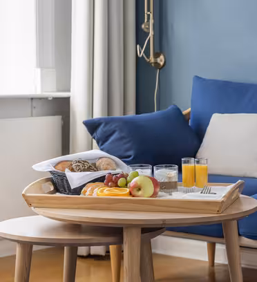 Ett mysigt vardagsrum har ett träbord med en frukostbricka med frukt, bröd, två glas apelsinjuice och vatten. Den blå soffan i bakgrunden har beige och blå kuddar, och naturligt ljus filtreras genom ett närliggande fönster.