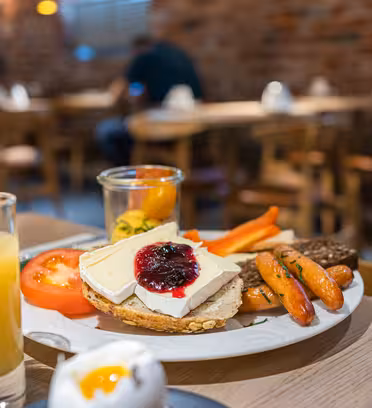 Ett träbord med en frukosttallrik med ost med sylt på bröd, körsbärstomater, morotsstavar och ett litet glas med apelsinjuice. Ett glas mjölk, ett glas juice och ett mjukkokt ägg finns bredvid. En caféinteriör syns i bakgrunden.