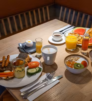 Ett träbord dukat för frukost med tallrikar med olika livsmedel inklusive frukt, yoghurt, bröd och bakverk. Drycker på bordet inkluderar kaffe, apelsinjuice och juicer. Besticken är snyggt arrangerade bredvid tallrikarna.