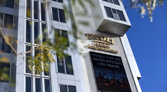 Hög byggnad med skylt "Tivoli Hotel & Congress Center", med en modern fasad med stora fönster. En digital skärm visar ett reklammeddelande. Trädgrenar ramar delvis in den nedre delen. Klarblå himmel i bakgrunden.