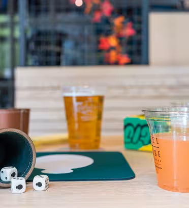 En bordsscen med två tärningar och en lädertärningskopp på en matta, två drinkar i plastmuggar – en gul och en rosa – och färgglada lådor i bakgrunden.