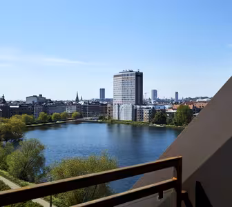 Utsikt från en balkong med utsikt över en sjö och en stadsbild. En modern, hög byggnad står framträdande vid sjökanten, omgiven av mindre byggnader. Himlen är klar och blå, med grönska längs vattnet.