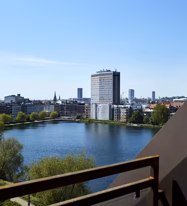 Utsikt från en balkong med utsikt över en sjö och en stadsbild. En modern, hög byggnad står framträdande vid sjökanten, omgiven av mindre byggnader. Himlen är klar och blå, med grönska längs vattnet.