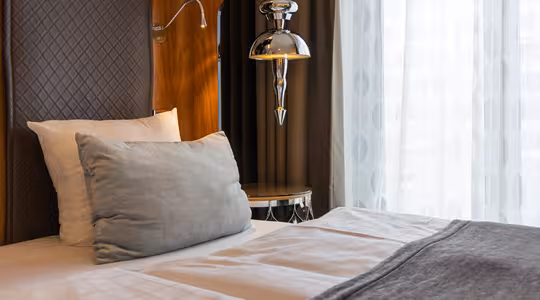 Ett mysigt hotellrum med en snyggt bäddad säng med vita och gråa sängkläder. En grå slängkudde läggs mot sänggaveln. En silverhängande lampa hänger ovanför ett litet runt sidobord bredvid sängen, nära ett fönster med skira gardiner.