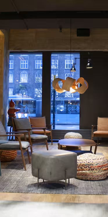 Modern hotellobby med mysig sittgrupp. Detta rum har en brun skinnsoffa, färgglada ottomaner och en dekorativ öppen spis. Stora fönster visar en urban gatubild. Varm belysning förstärker den inbjudande atmosfären.