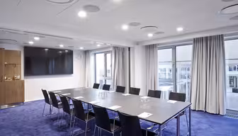 Ett modernt konferensrum med ett långt bord omgivet av svarta stolar på en blå matta. Papper och glas vatten ställs på bordet. Rummet har stora fönster, en väggmonterad TV och infälld belysning.