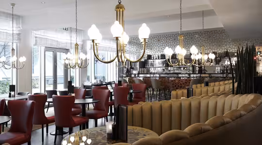 En elegant restauranginteriör med eleganta ljuskronor som lyser upp utrymmet. Röda stolar omger runda bord och en svängd beige soffa ger sittplatser. Väggarna har geometriska mönster och stora fönster släpper in naturligt ljus.
