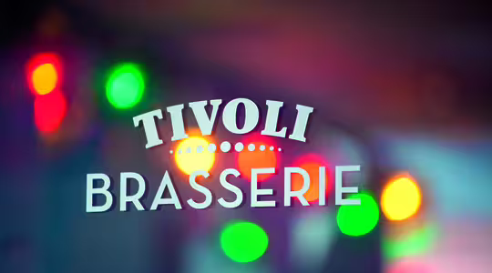Bilden visar ett glasfönster med texten "TIVOLI BRASSERIE" med vita bokstäver. I bakgrunden syns färgglada suddiga ljus, vilket skapar en levande och festlig atmosfär.