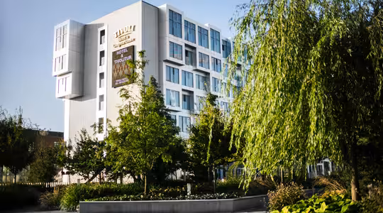 Modern vit hotellbyggnad med stora fönster, omgiven av grönskande träd och anlagda trädgårdar. Hotellets namn och annons syns på utsidan. Klarblå himmel i bakgrunden.